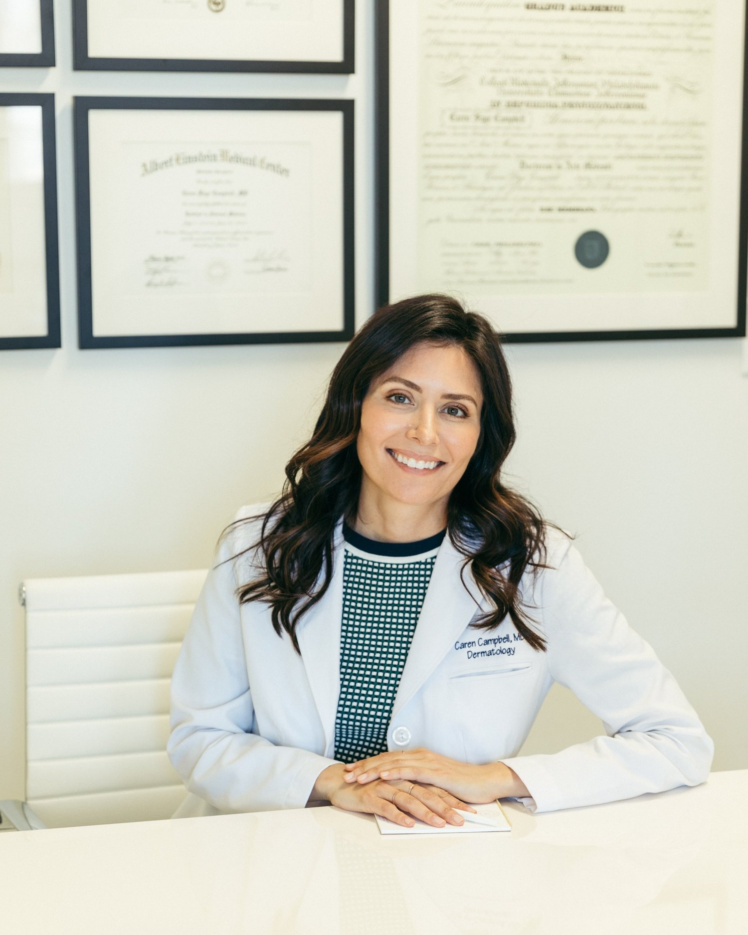 Dr. Caren Campbell, MD, a board-certified dermatologist, smiling in her office with framed medical diplomas in the background. Expert in skincare, acne treatments, and skin health.