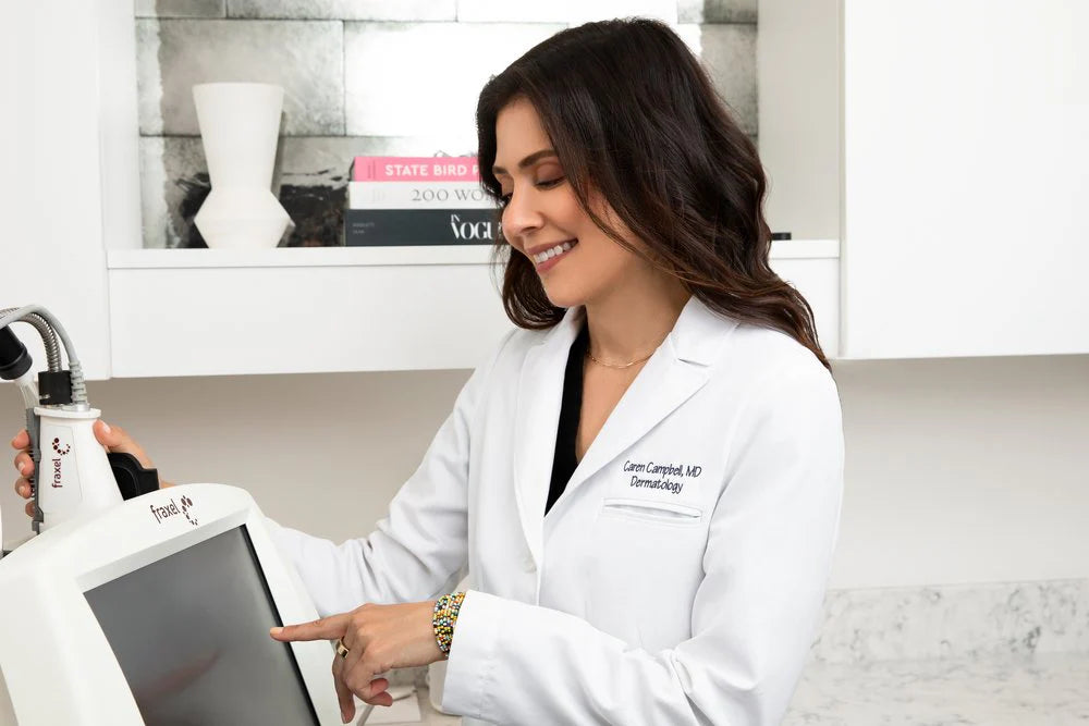 Dr. Caren Campbell in her dermatology office, wearing a white coat and demonstrating the Fraxel machine, a state-of-the-art device used for skin resurfacing and rejuvenation treatments.